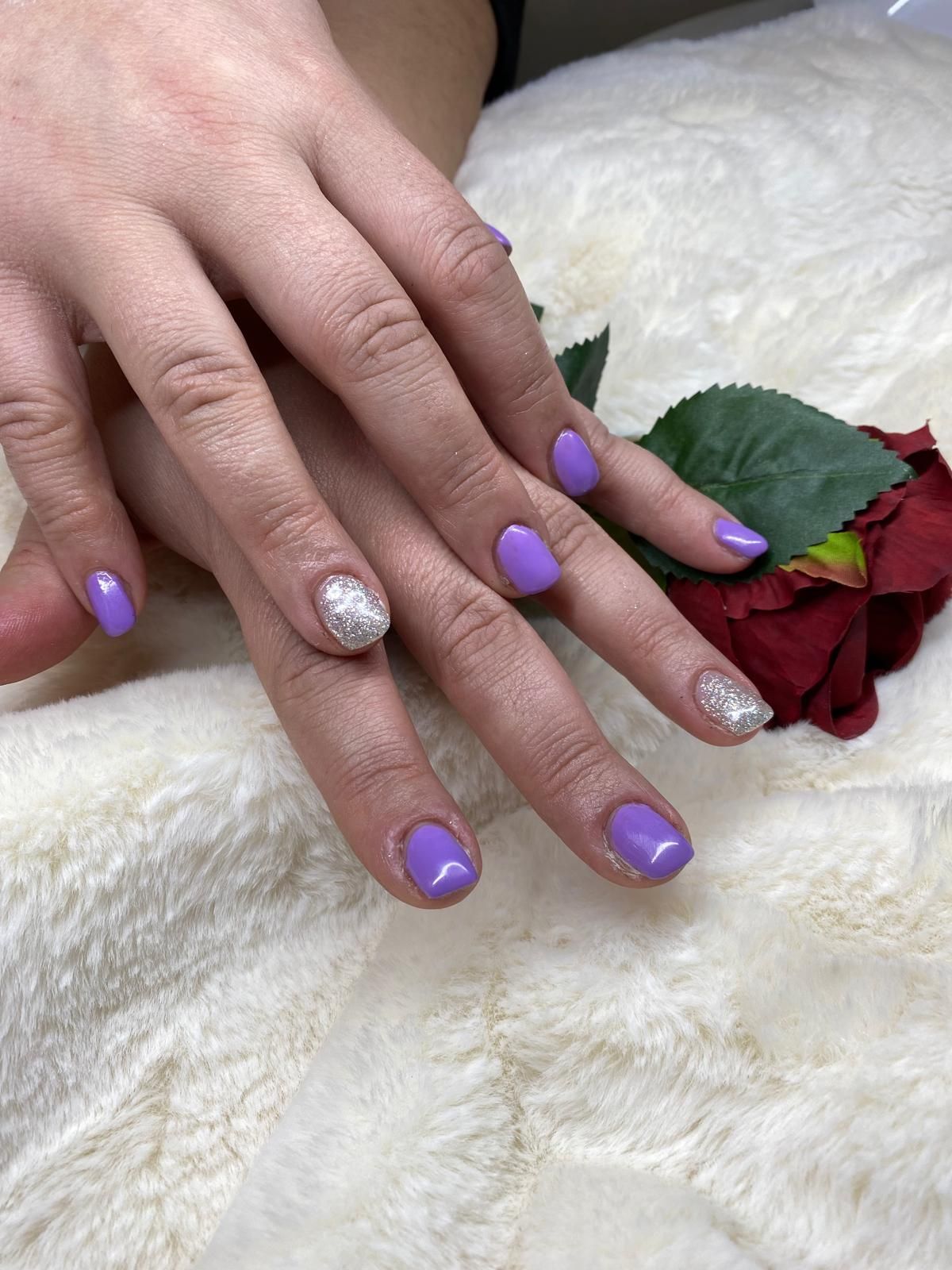 Hände mit lavendelfarbenem Nagellack und silbernen Glitzerakzenten auf einem flauschigen Untergrund mit einer roten Rose.
