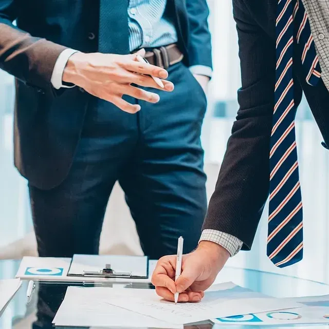 Dos profesionales de negocios con traje revisan documentos; uno escribe y el otro gesticula.