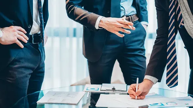 Tres hombres de negocios vestidos de traje discuten documentos en una mesa de cristal; uno de ellos firma el papeleo.