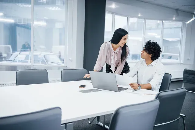 Dos mujeres en una oficina, una de pie e inclinada, la otra sentada, mirando una computadora portátil y papeles sobre una mesa blanca.
