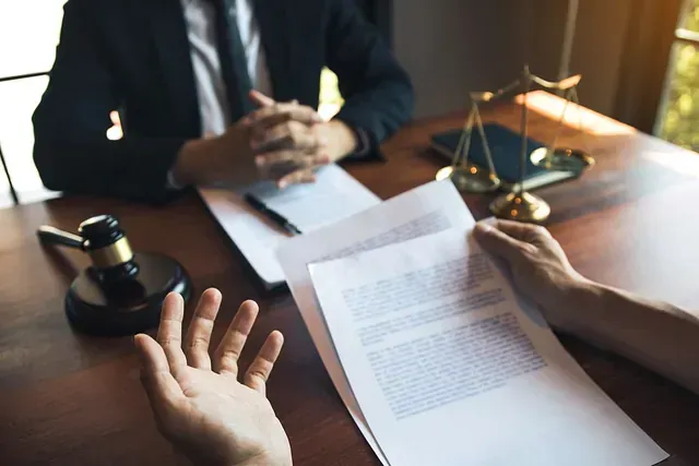 Una persona con documentos gesticula mientras habla con un abogado de traje, sentado en un escritorio. Escena de tribunal, balanza de la justicia y mazo.