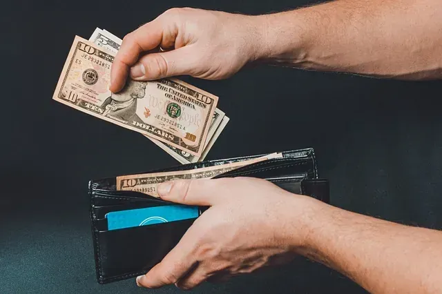 Manos colocando varios billetes de diez dólares estadounidenses en una billetera negra sobre un fondo oscuro.