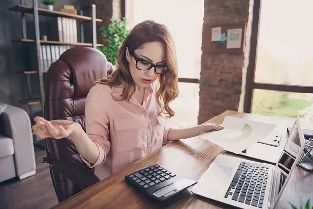 Mujer con gafas, con aspecto perplejo, sentada en un escritorio con calculadora, ordenador portátil y papeles en un entorno de oficina.