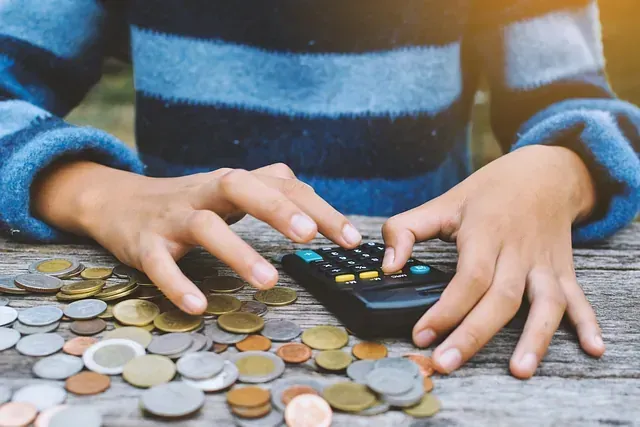 Manos utilizando una calculadora, rodeadas de pilas de monedas sobre una superficie de madera.