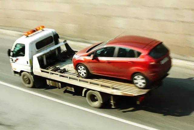 Un coche rojo remolcado por un camión de plataforma circula por la carretera. El camión es blanco y el entorno parece ser exterior.