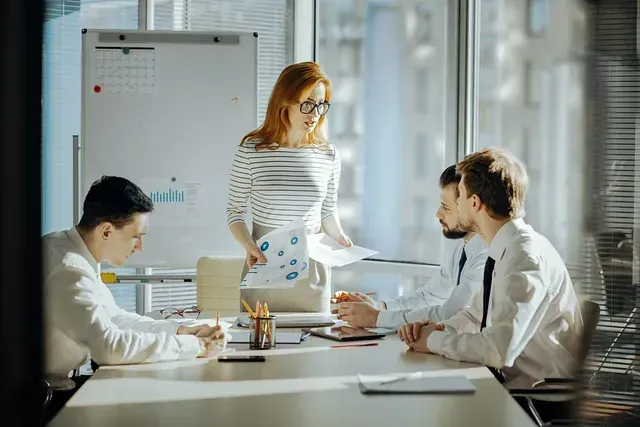 Una mujer con camisa a rayas dirige una reunión en una oficina luminosa. Tres hombres están sentados a una mesa, revisando documentos.