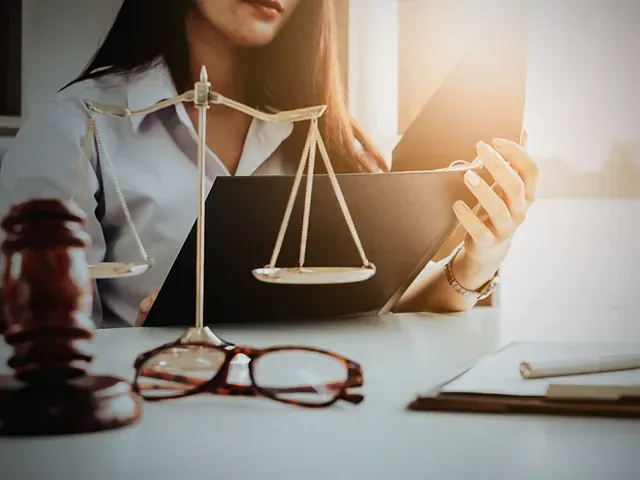 Una persona sosteniendo documentos, con un mazo, una balanza y gafas sobre un escritorio.