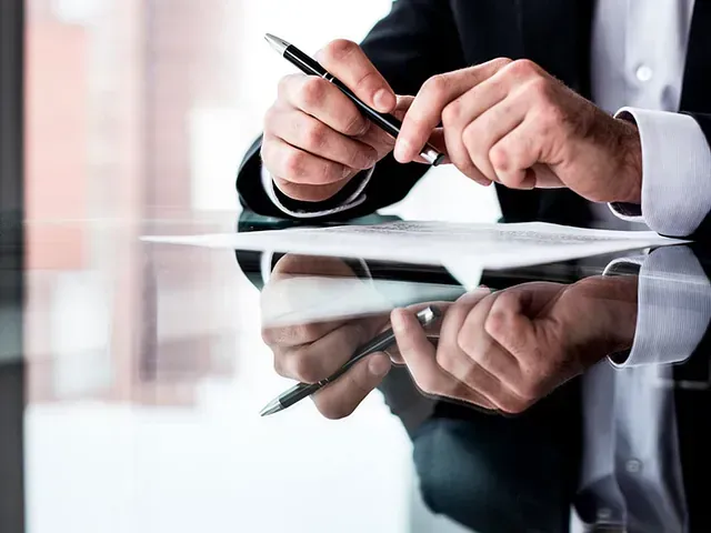 Una persona con traje sostiene un bolígrafo sobre un documento, con las manos reflejadas en una mesa de cristal.