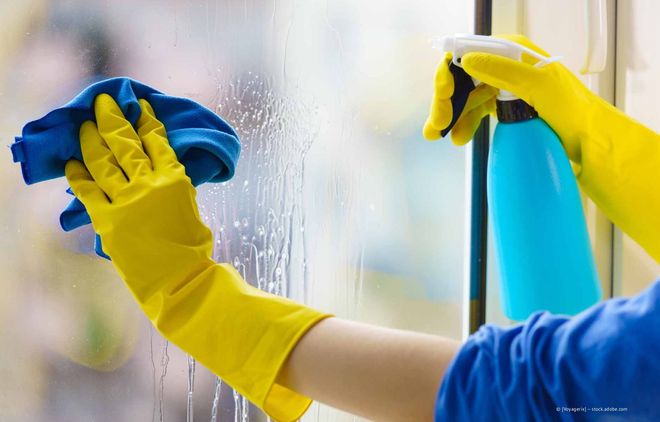 Hände in gelben Handschuhen wischen ein Fenster mit blauen Putztüchern ab.