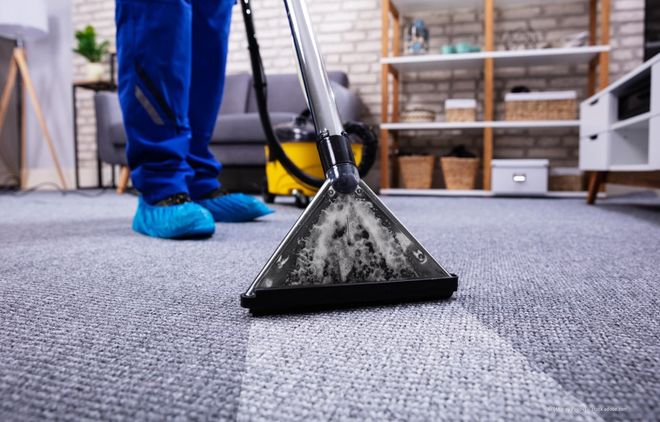 Ein Teppichreiniger saugt einen grauen Teppich in einem Wohnhaus; in der Nähe befindet sich eine Person in blauer Hose und Schuhen.