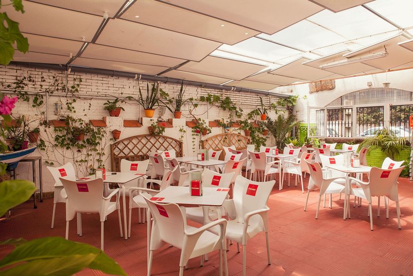 Un patio exterior muy iluminado, con filas de mesas y sillas blancas sobre un suelo rojo.