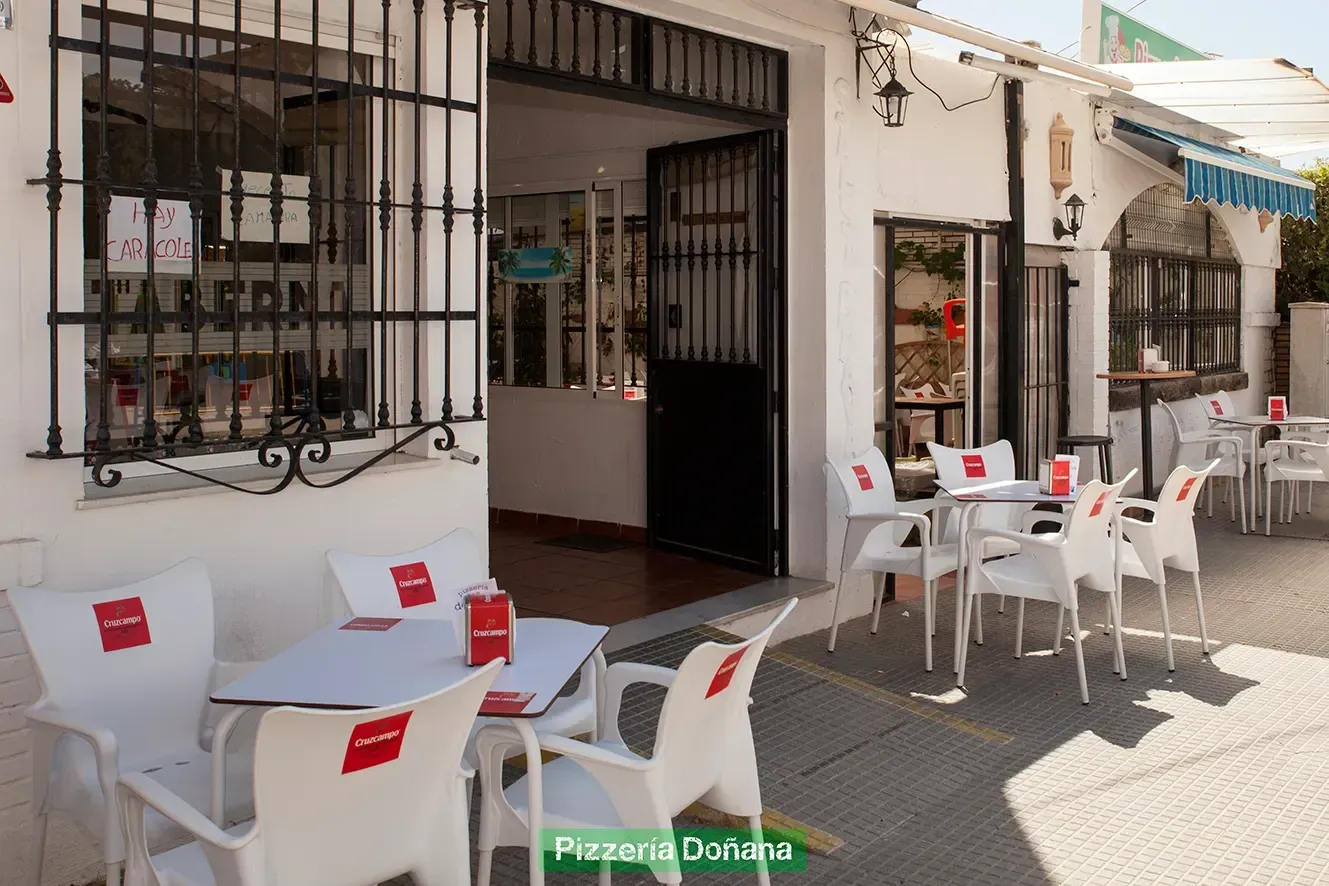 Una terraza de restaurante con mesas al aire libre y sillas blancas bajo un toldo azul en una calle soleada.