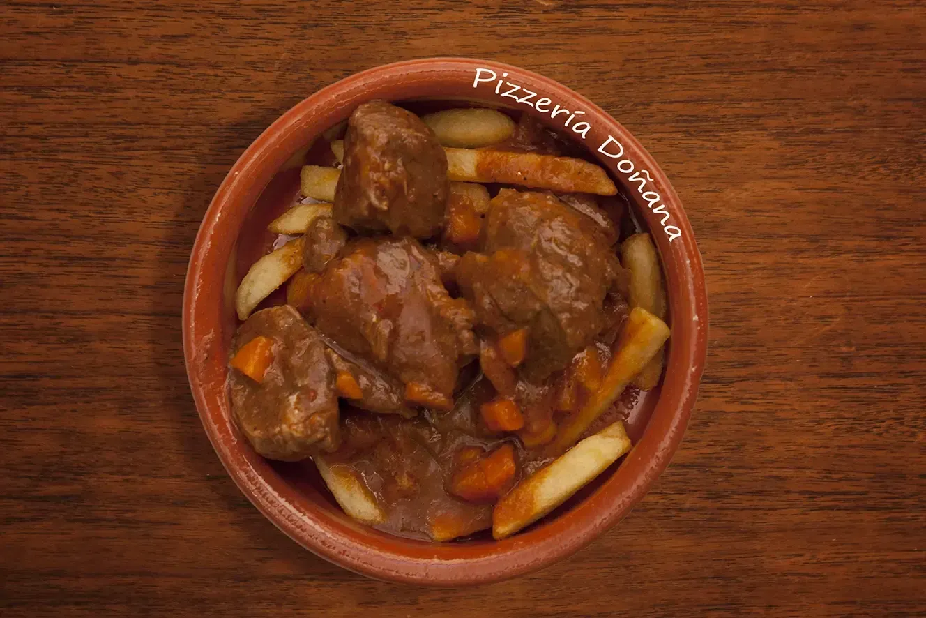 Un cuenco de barro lleno de estofado de ternera y zanahorias servido sobre patatas fritas en una mesa de madera.
