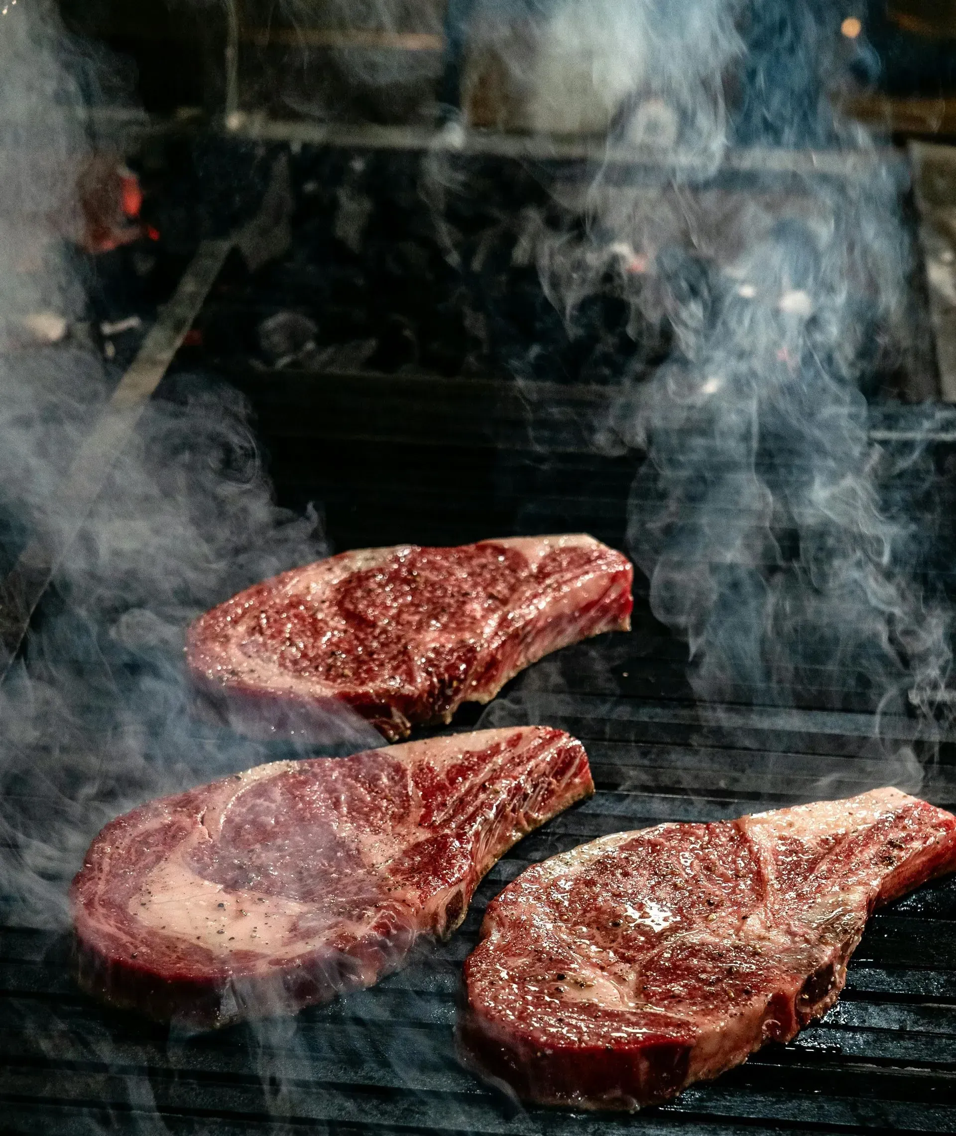 Tres filetes de chuletón crudos y veteados, asándose a la parrilla sobre una rejilla caliente, con humo que se eleva.