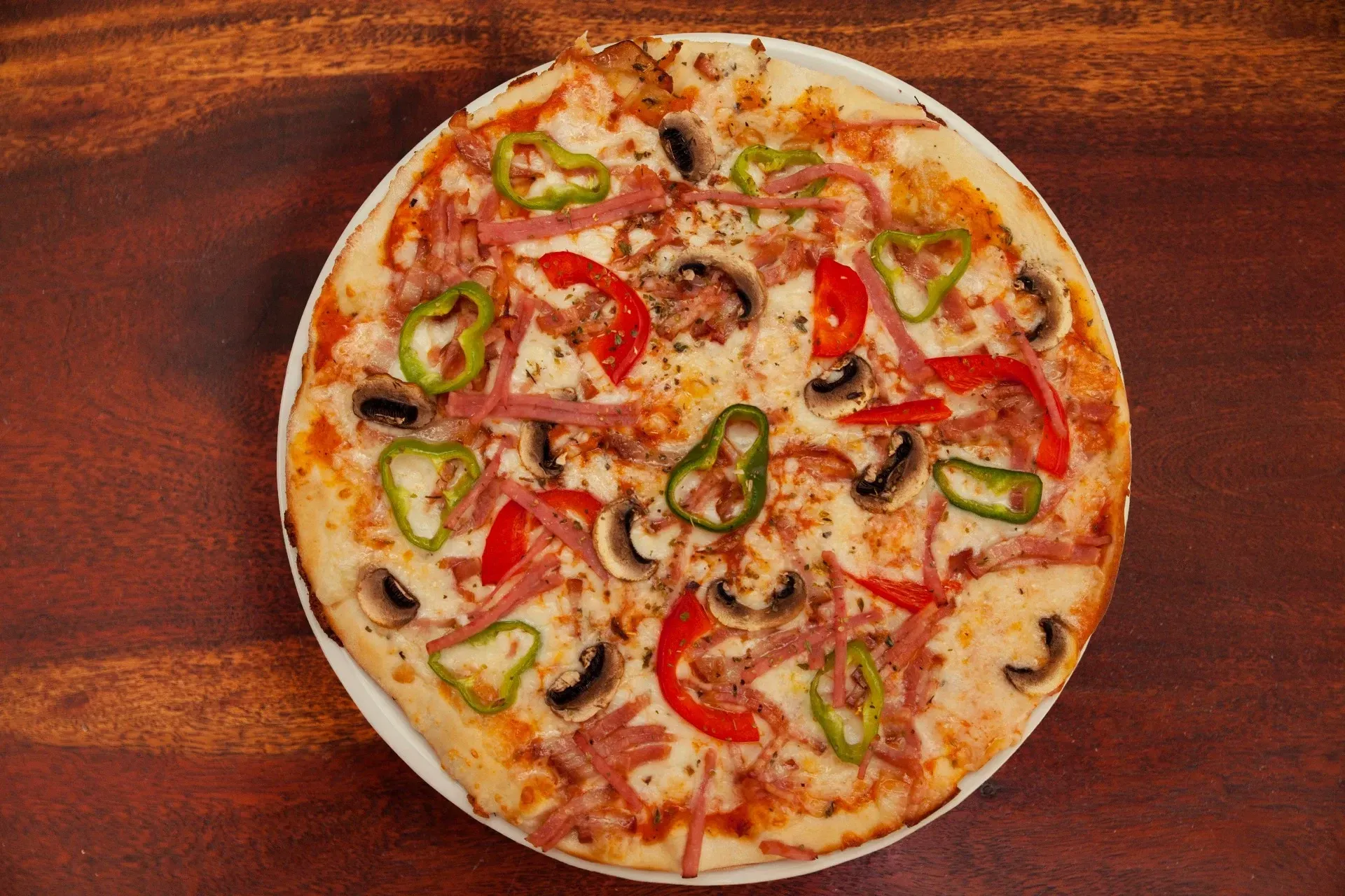 Una pizza cubierta con jamón, champiñones y pimientos reposa sobre un plato blanco con un fondo de madera oscura.