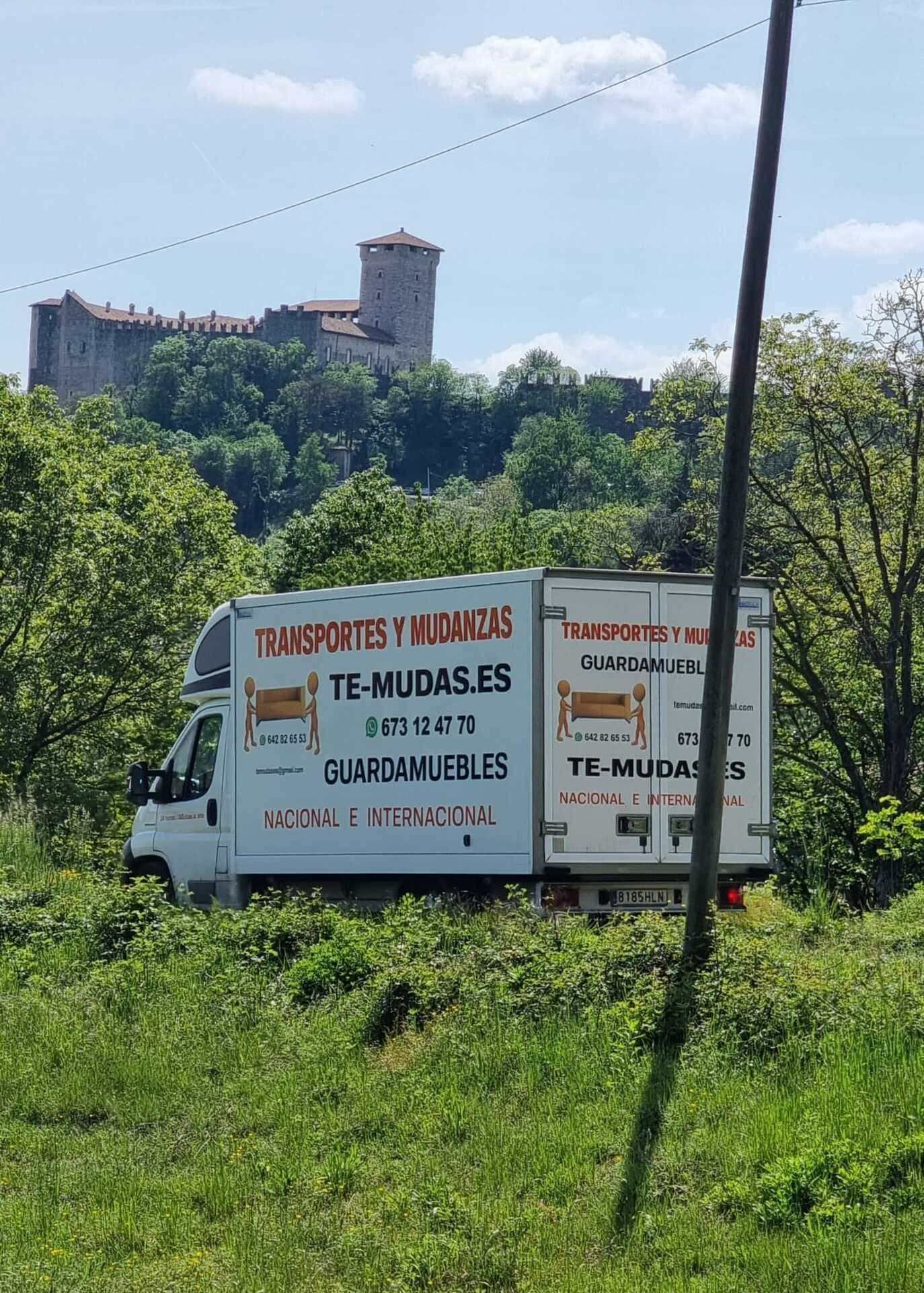 Camioneta blanca en un campo verde con un castillo en la cima de una colina al fondo.