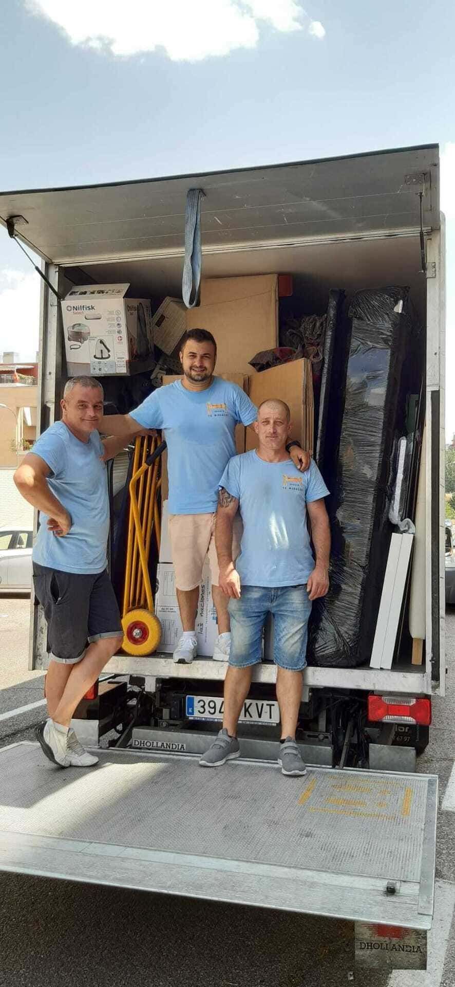 Tres personas de pie en la parte trasera de un camión de mudanzas cargado de muebles y equipos.