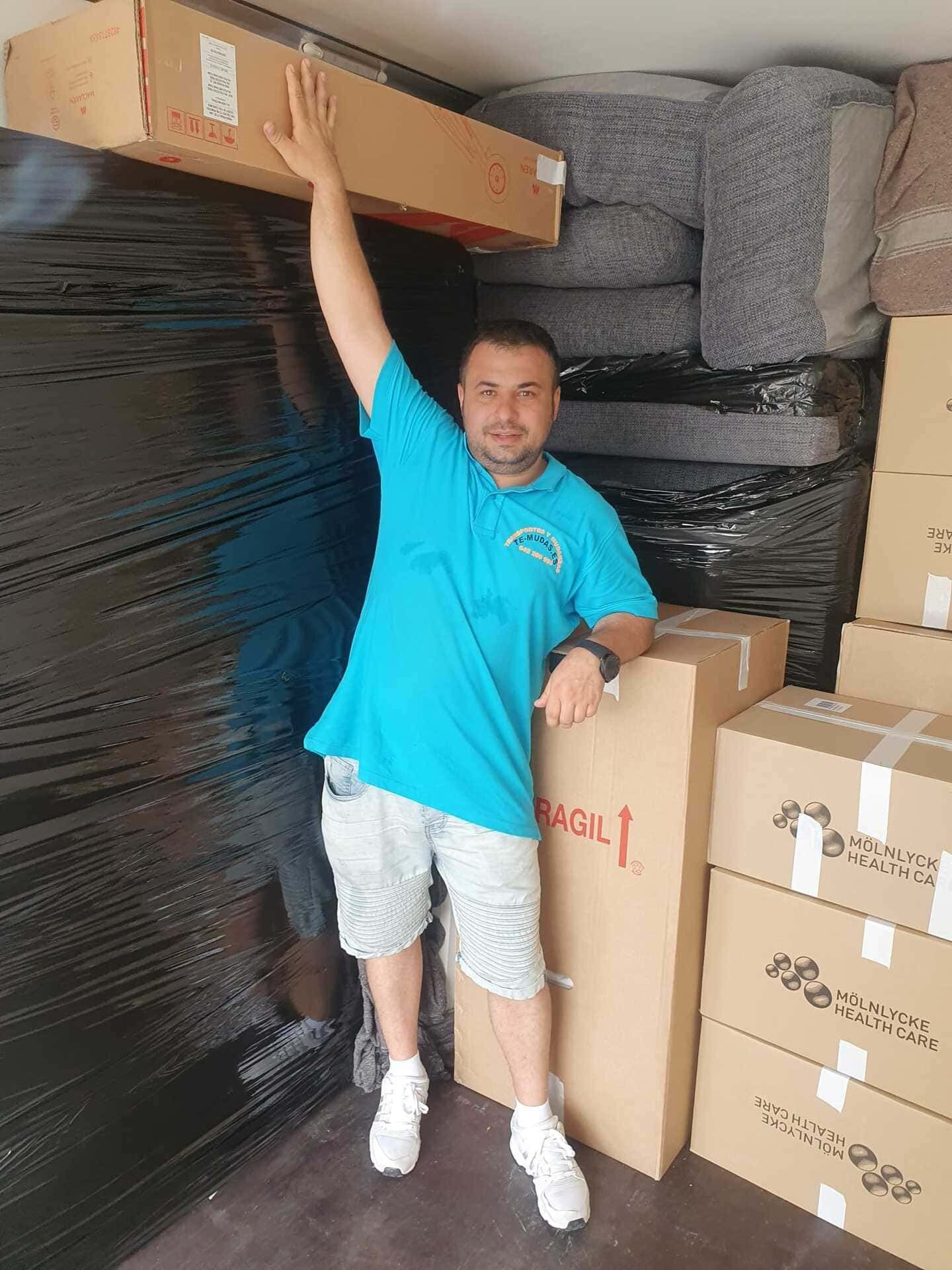 Un hombre posa en un almacén junto a cajas apiladas, extendiendo el brazo para sujetar una caja de cartón larga.