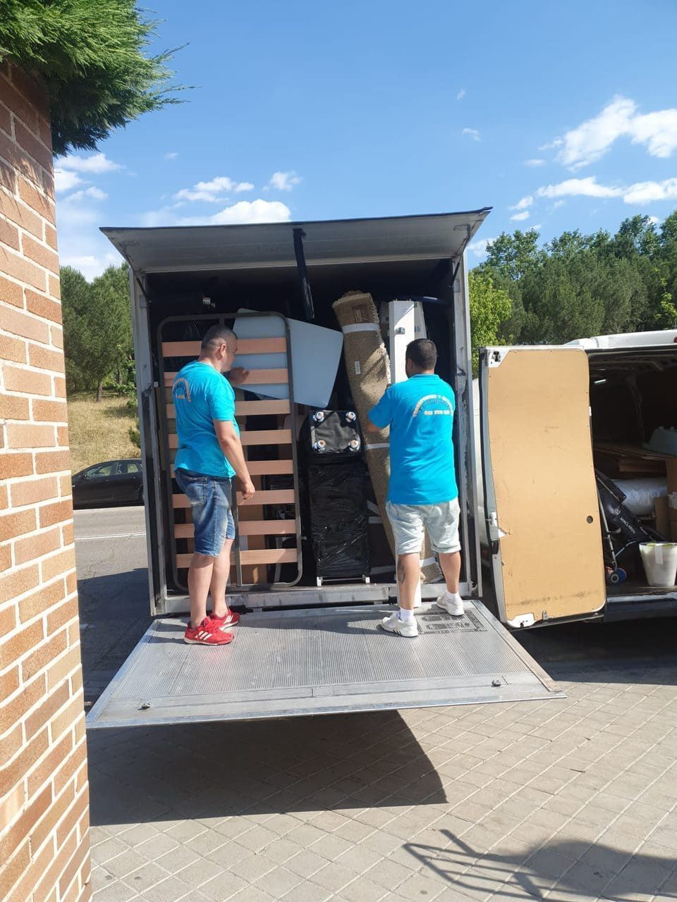 Dos operarios de mudanzas cargan cajas apiladas en la rampa de un camión bajo un cielo azul.