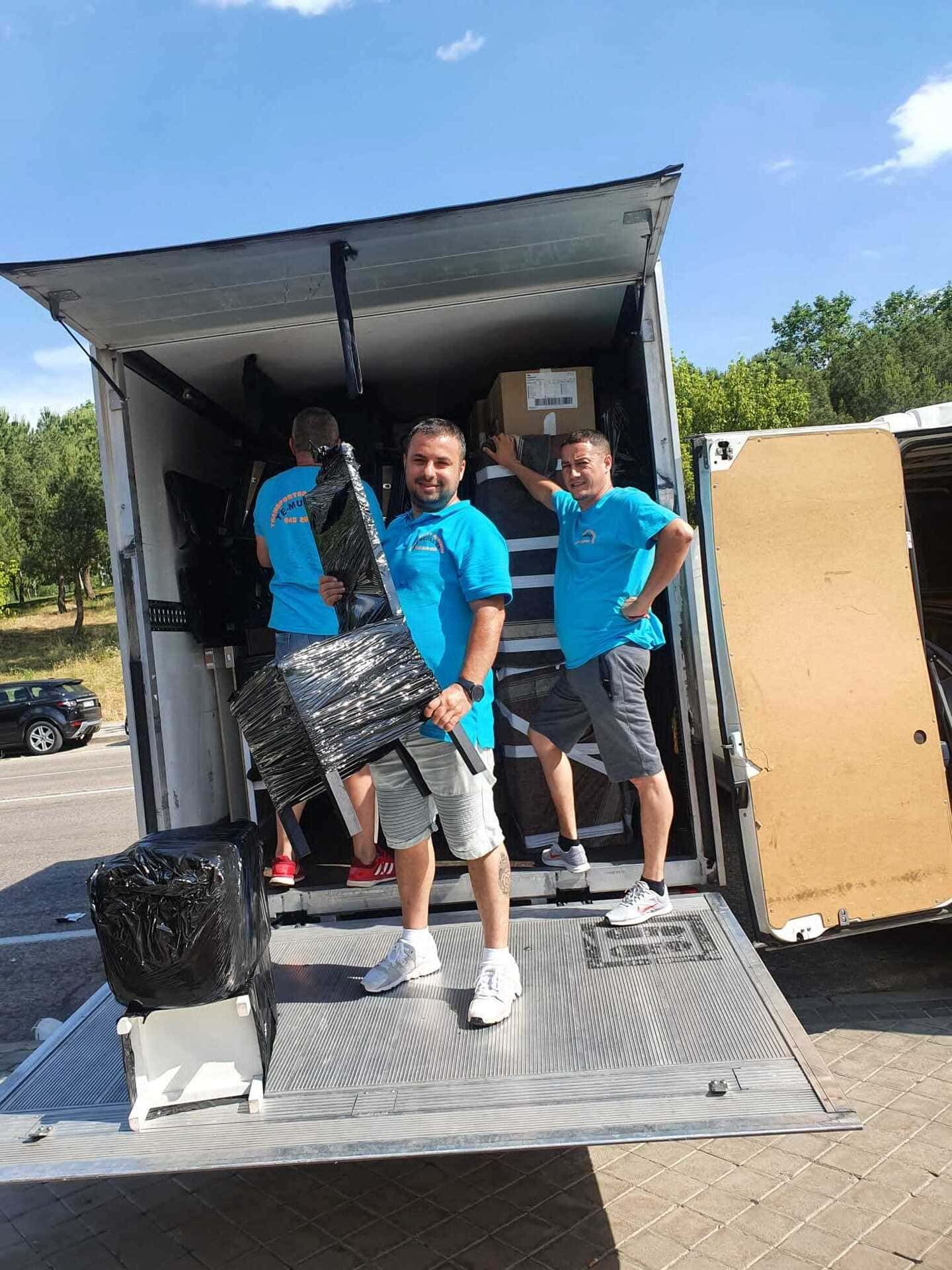 Tres personas con camisas azules están de pie en la parte trasera de un camión de mudanzas cargado de muebles y cajas.