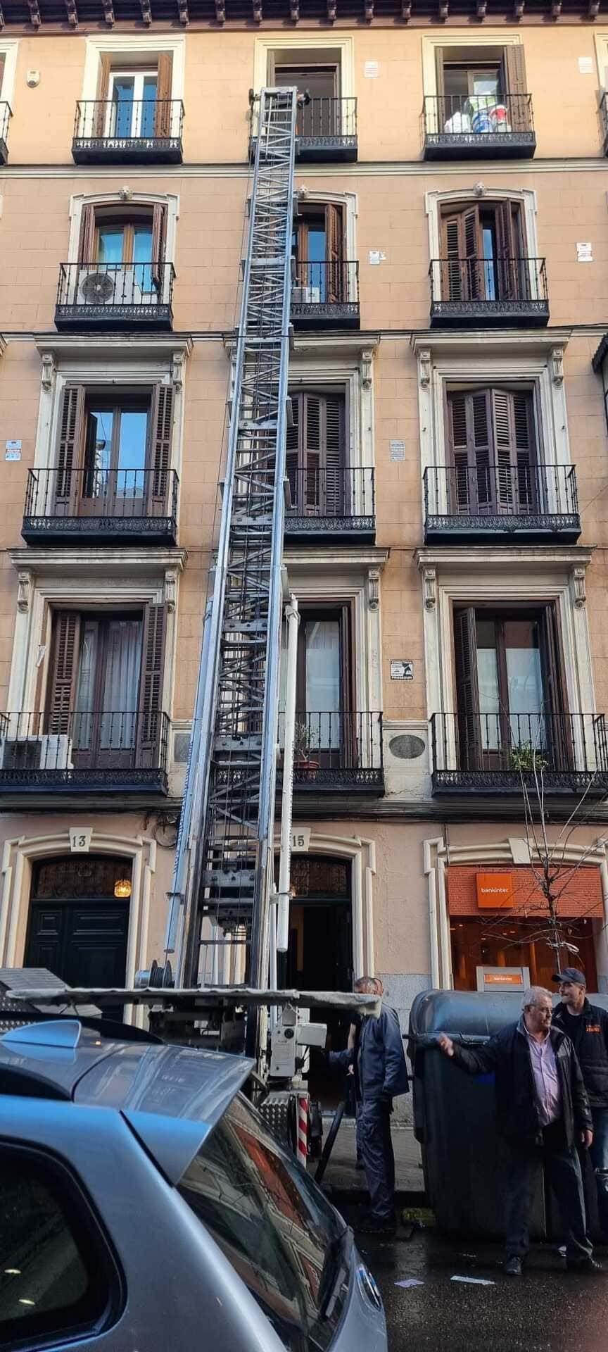 Fachada de un antiguo edificio de apartamentos con balcones, una escalera de incendios metálica y tiendas a pie de calle detrás de coches aparcados.