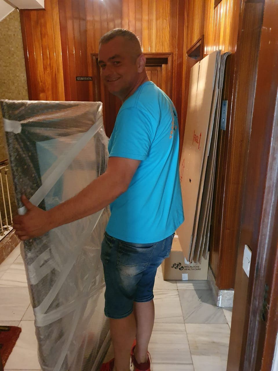 Un hombre transporta un colchón envuelto a través de un pasillo con paneles de madera.