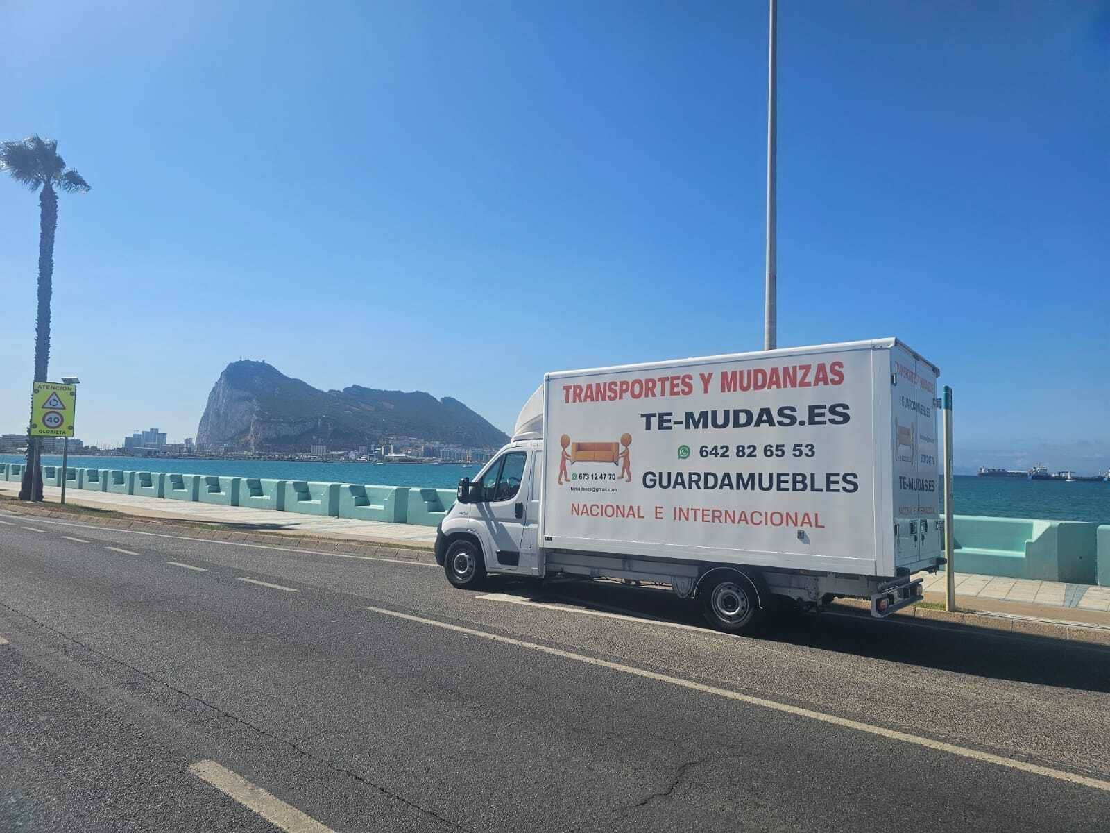 Furgoneta de reparto blanca en una carretera costera junto a aguas turquesas y una isla rocosa bajo un cielo azul brillante.