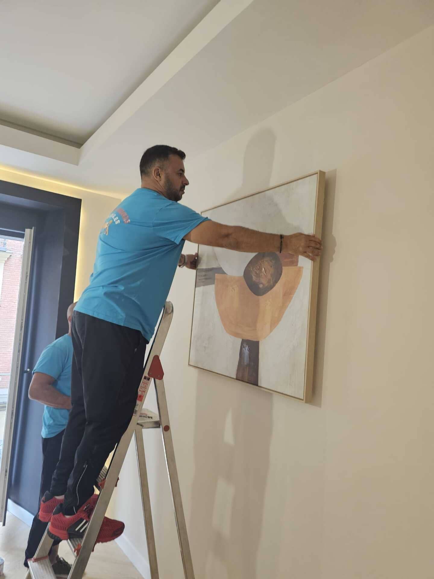 Dos hombres cuelgan un cuadro enmarcado en una pared beige usando una escalera.