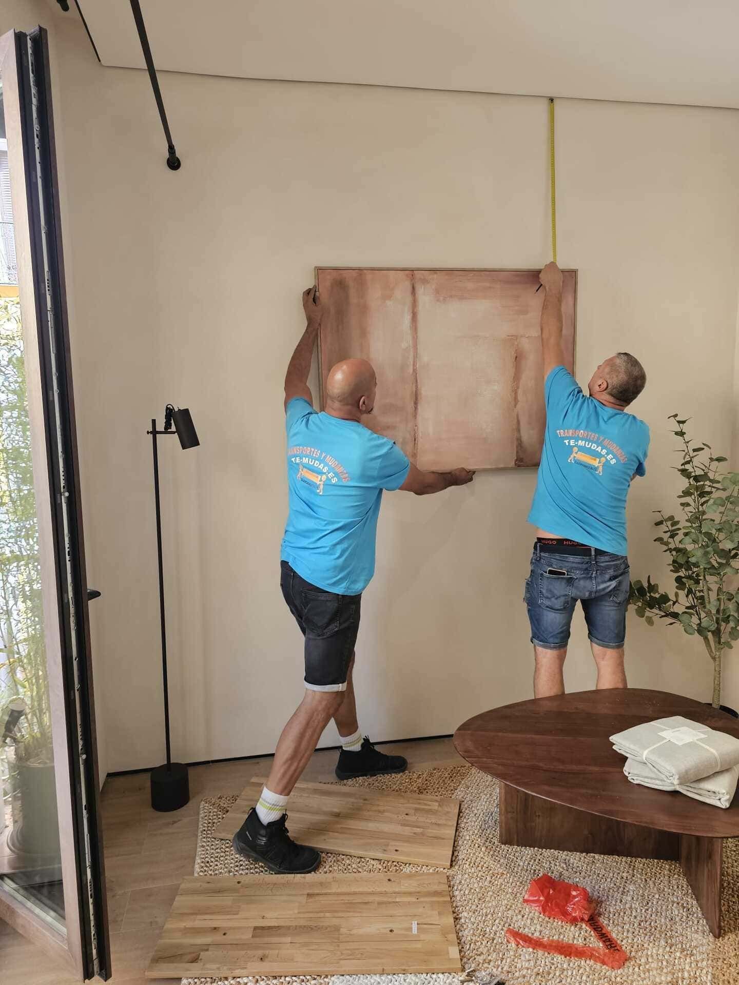 Dos trabajadores con camisas azules levantan un gran panel de madera que queda pegado a la pared en una habitación con una mesa redonda y plantas.