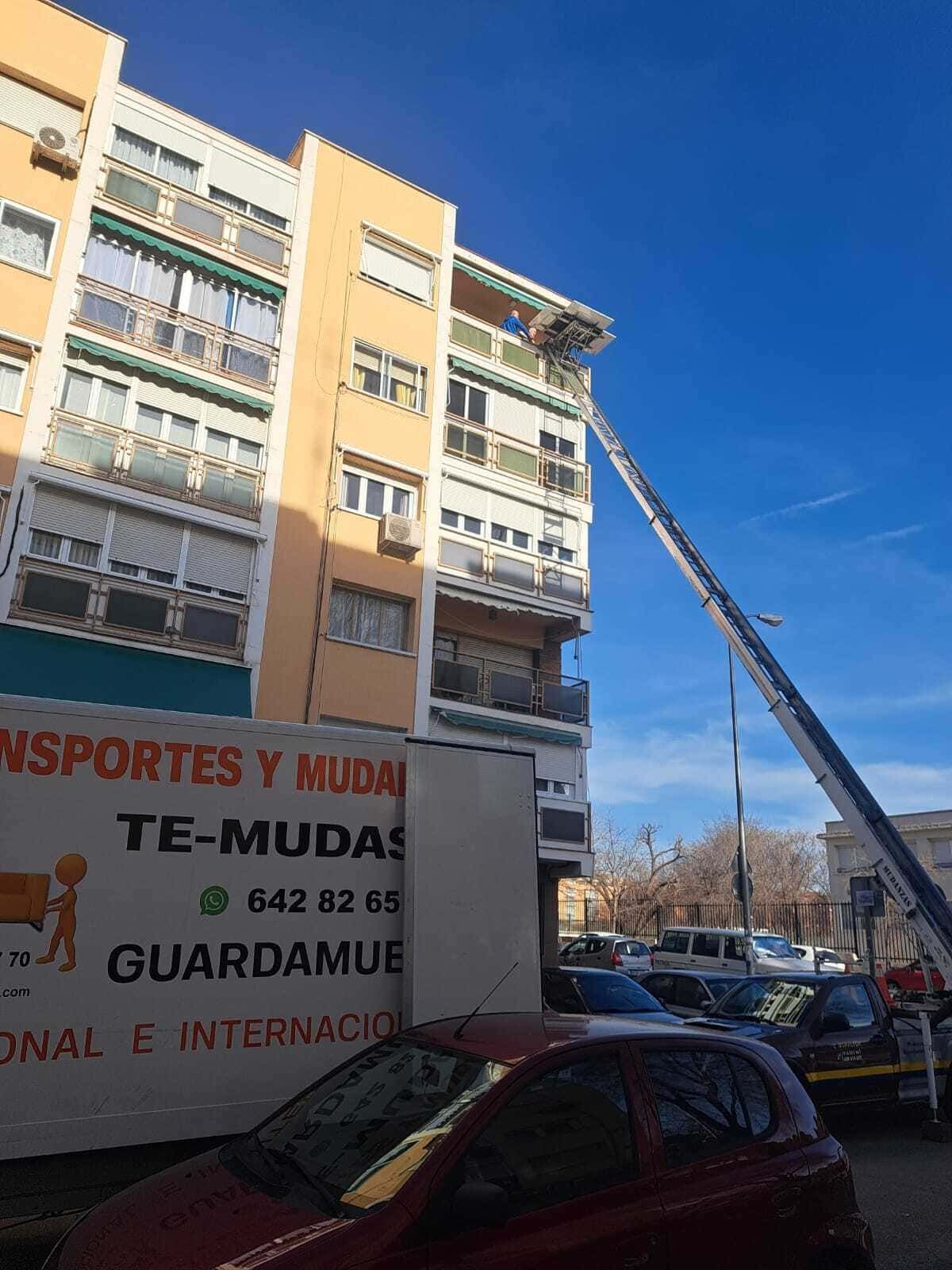 Camión de mudanzas subiendo muebles al balcón de un apartamento junto a un edificio beige en una calle soleada.