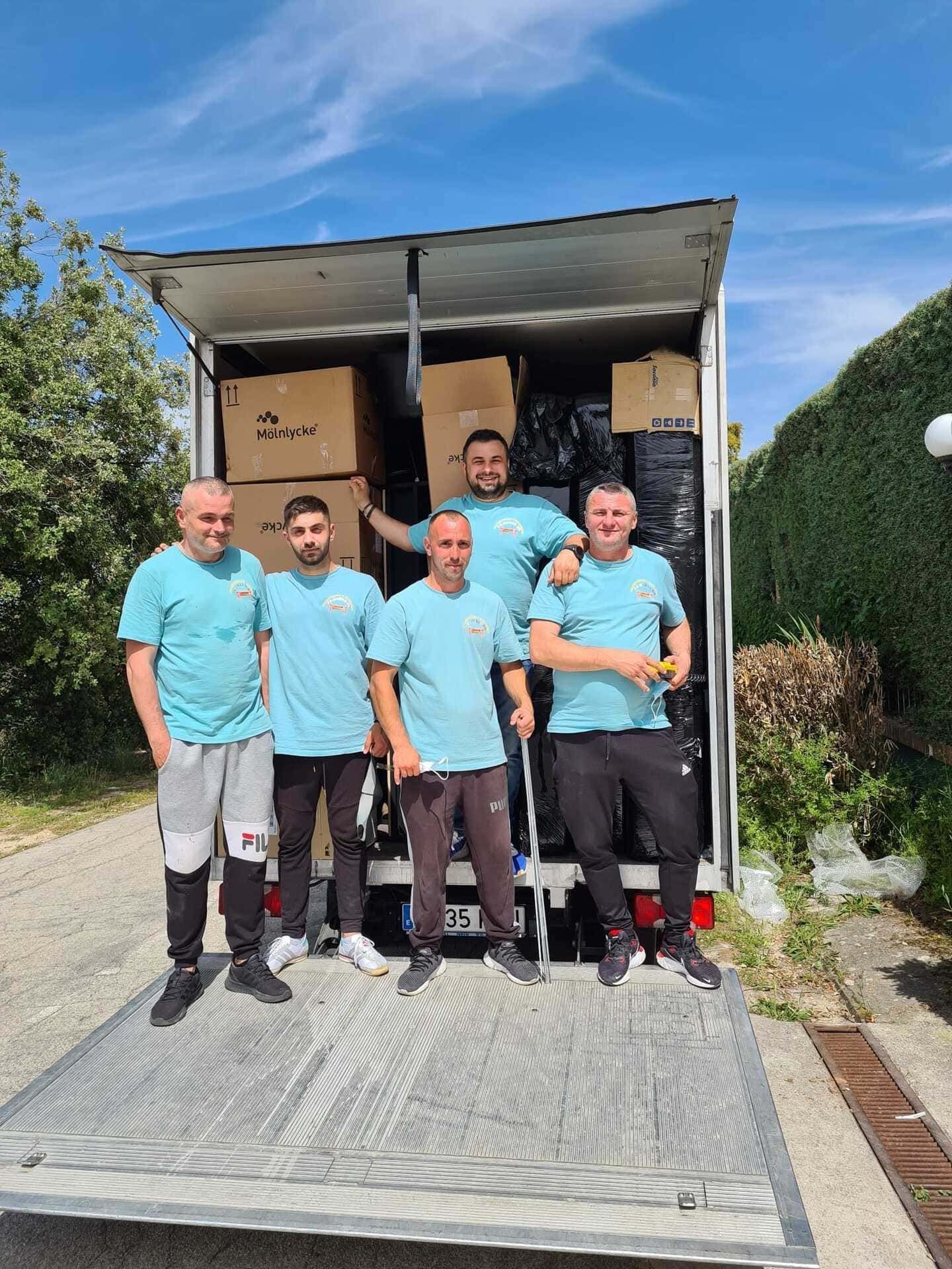 Cinco trabajadores con camisas turquesa están de pie en un camión de mudanzas con cajas en la parte trasera.