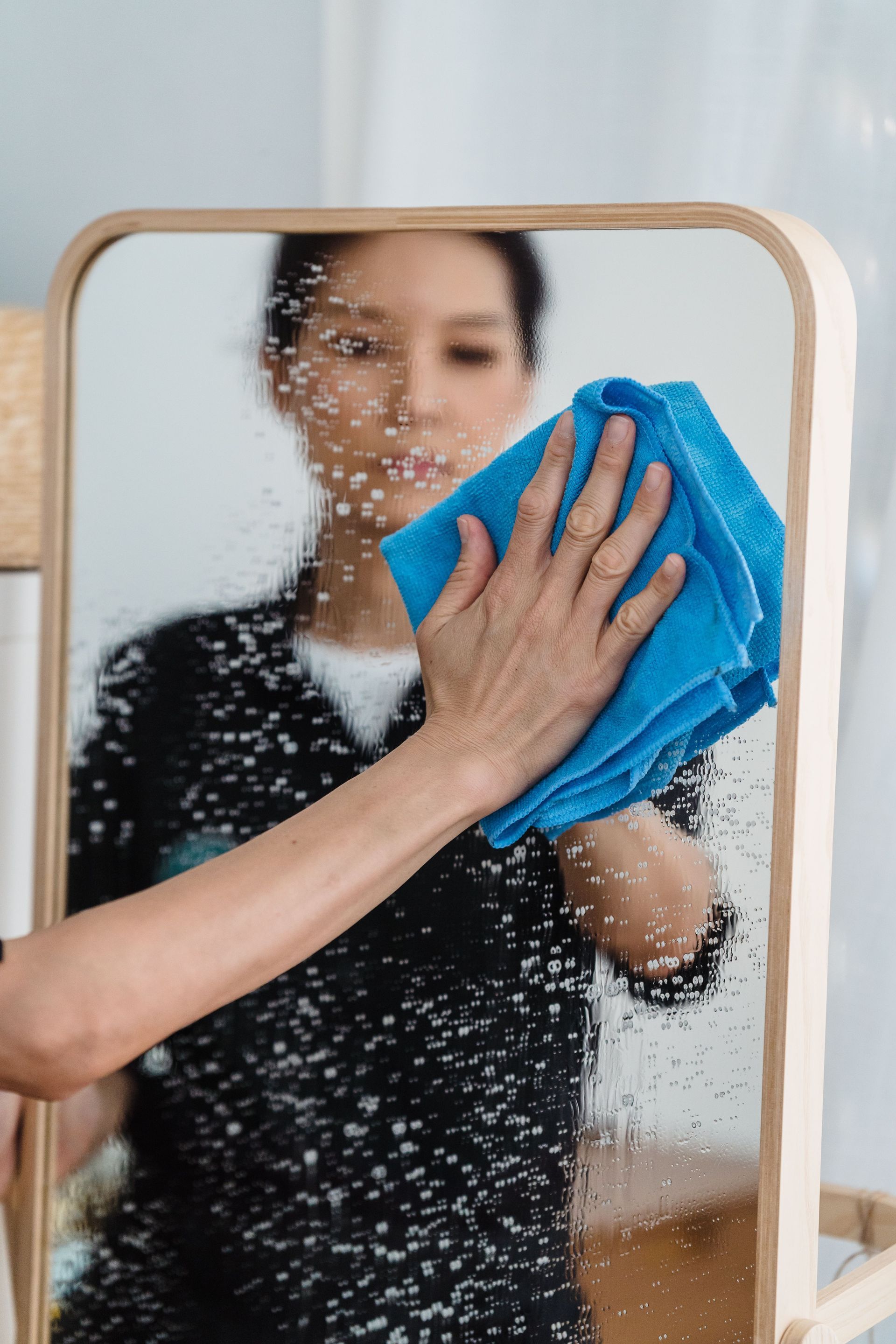 Una persona limpia un espejo con un paño azul, eliminando manchas de agua en una habitación.