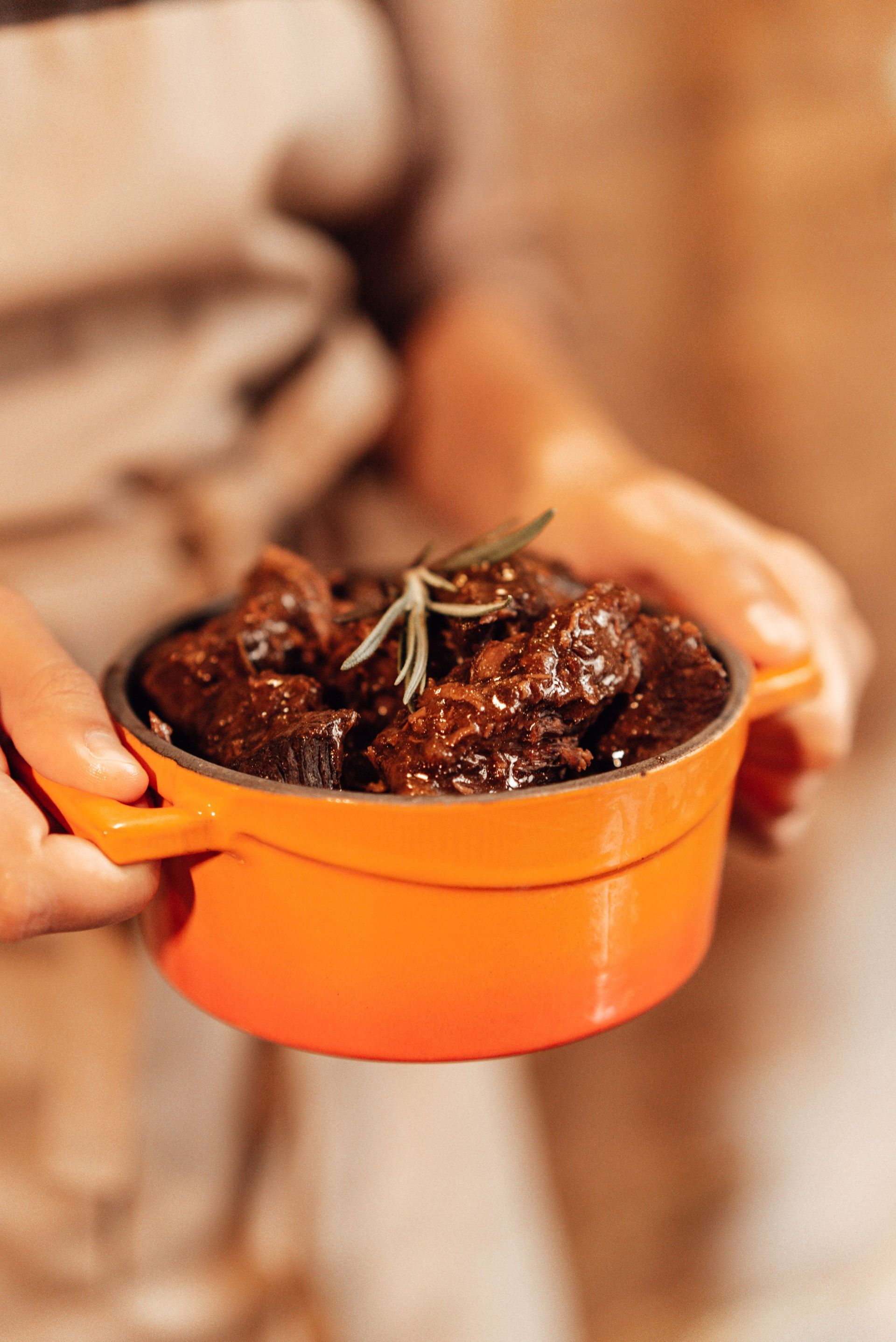 Des mains tiennent un pot orange rempli de terre, avec un petit brin de romarin