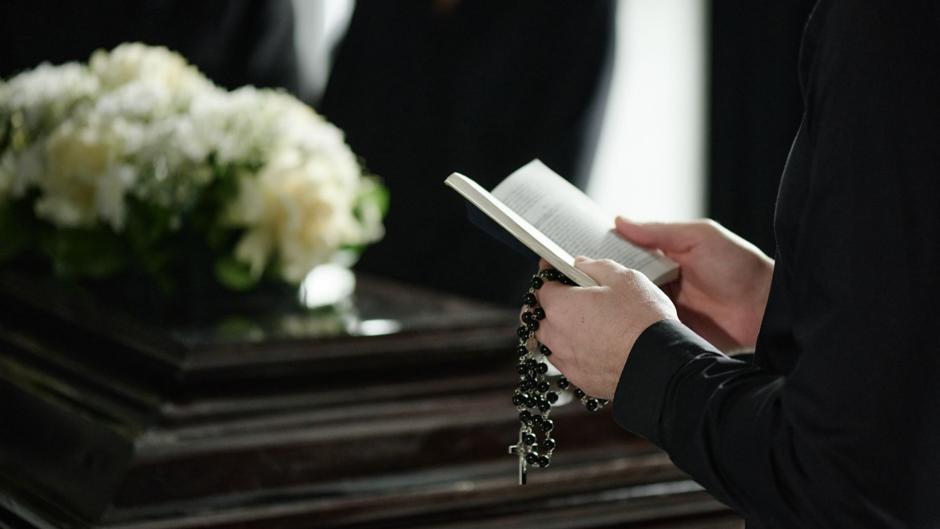 Une personne tenant un livre et un chapelet devant un cercueil.