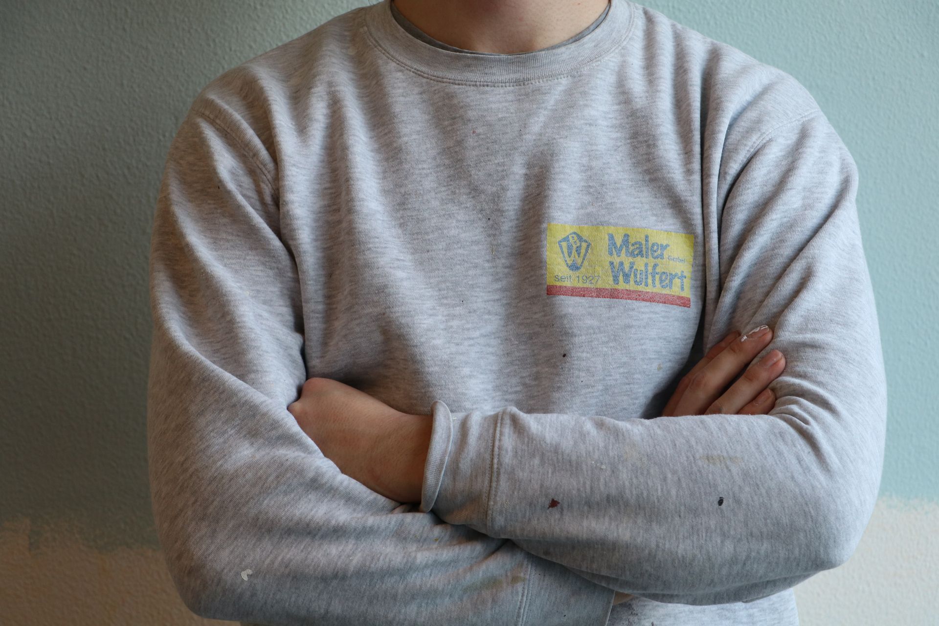 Ein Mann in einem grauen Pullover mit einem Namensschild steht mit verschränkten Armen da.