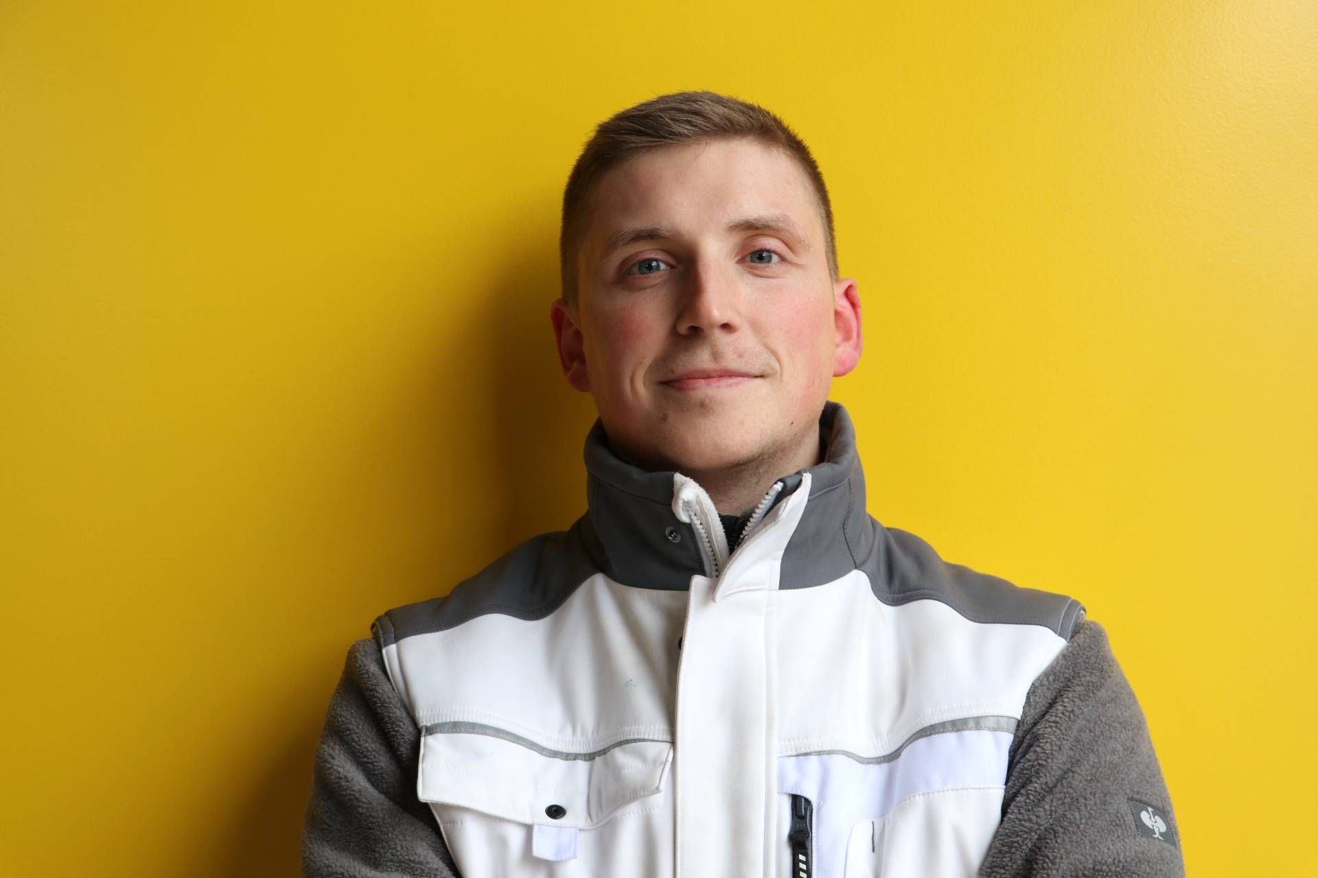 Ein Mann in einer weiß-grauen Jacke steht vor einer gelben Wand.