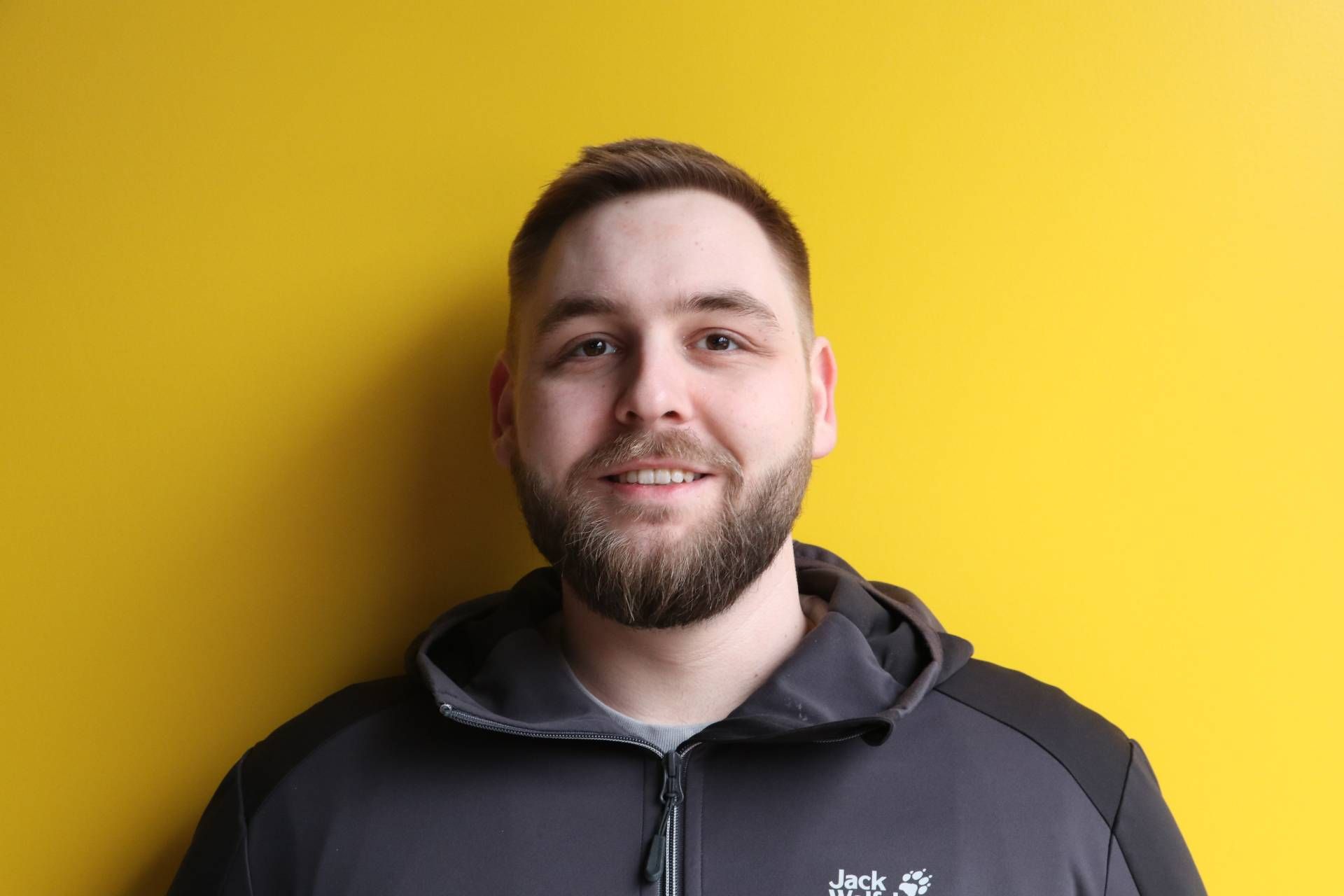 Ein Mann mit Bart lächelt vor einer gelben Wand.