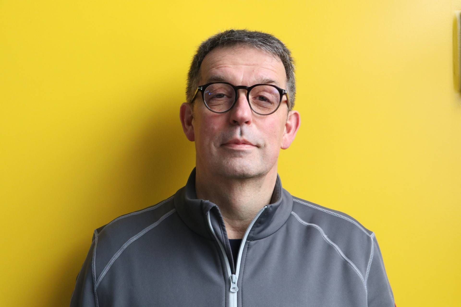 Ein Mann mit Brille und grauer Jacke steht vor einer gelben Wand.