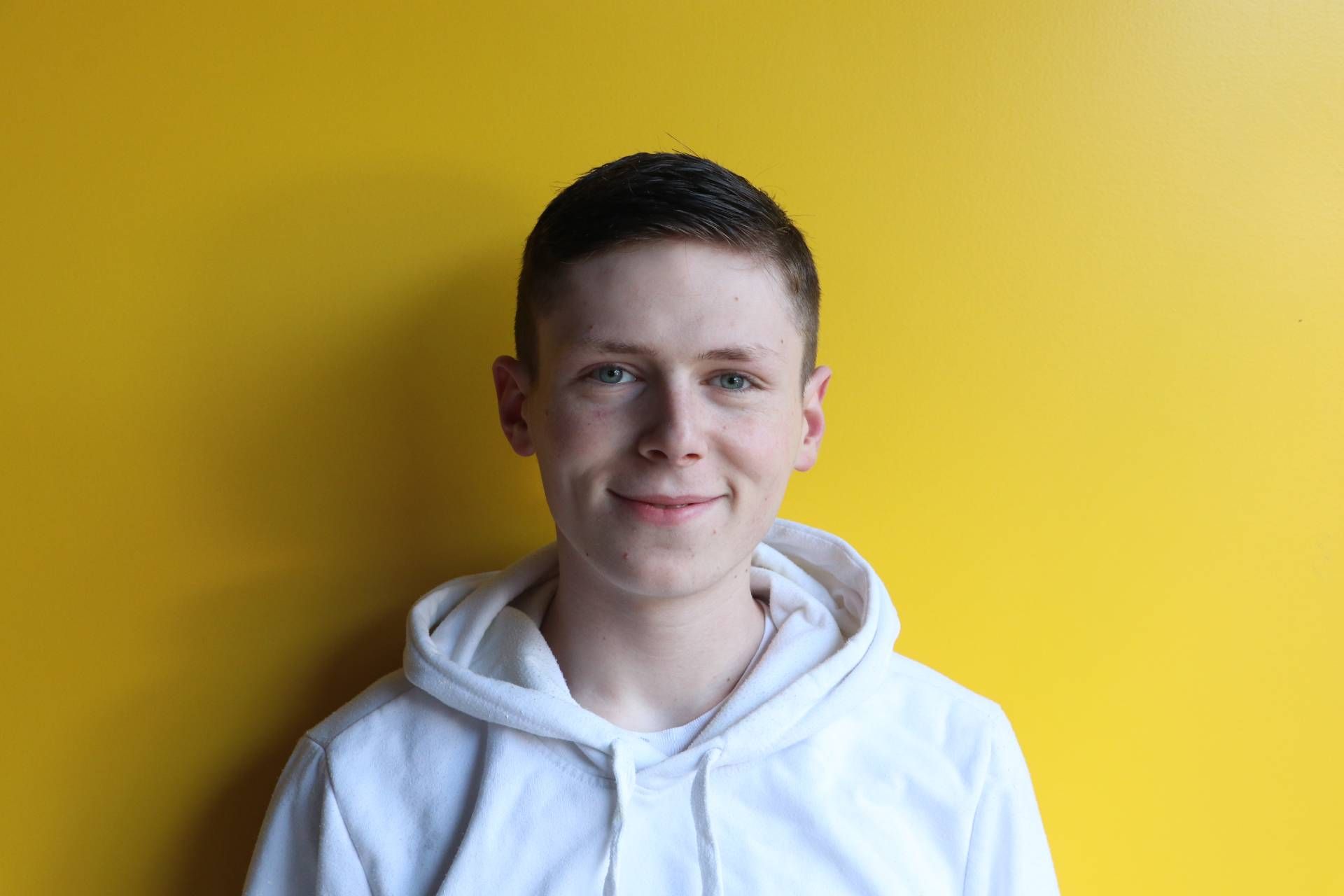 Ein junger Mann in einem weißen Kapuzenpullover steht vor einer gelben Wand.