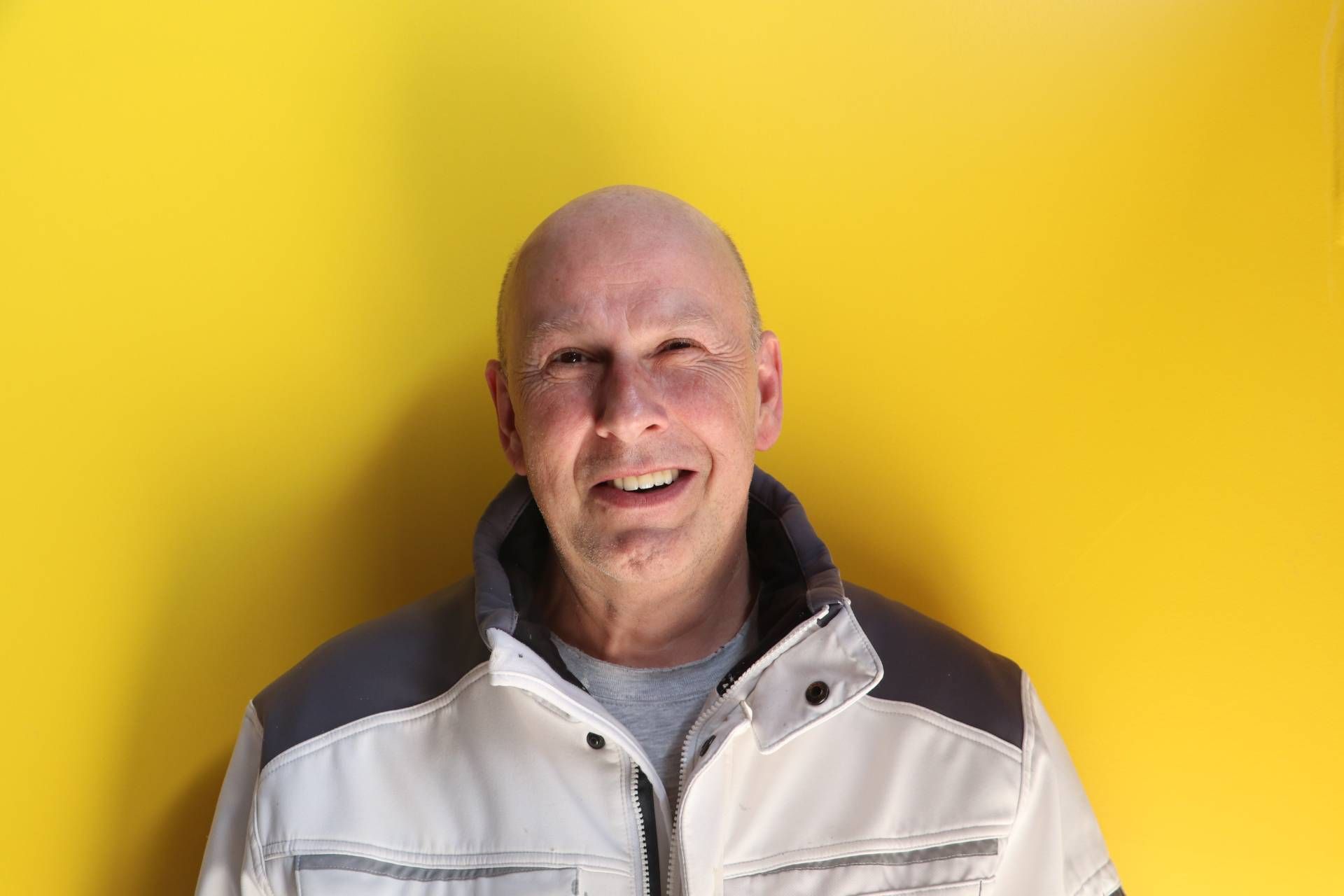 Ein kahlköpfiger Mann in einer weißen Jacke lächelt vor einer gelben Wand.