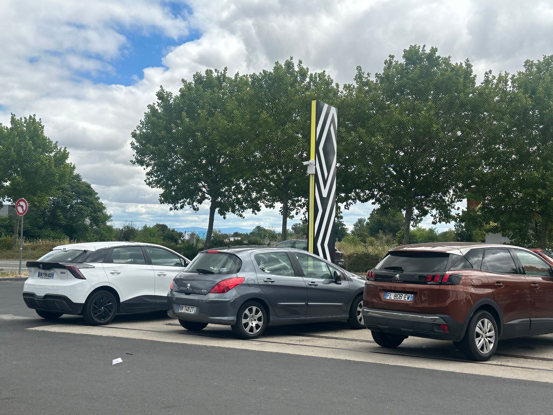 Trois voitures garées sur un parking goudronné, devant des arbres et un grand poteau rayé noir et blanc.