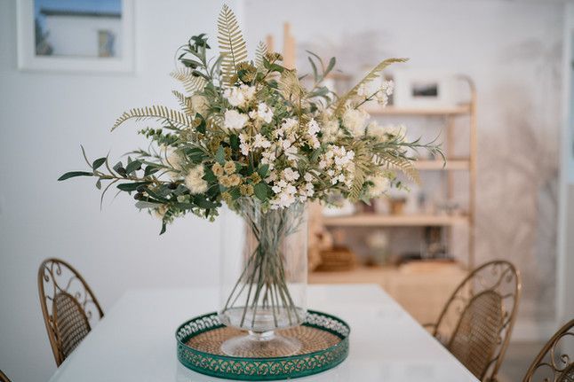 Arreglo floral en jarrón de vidrio sobre mesa; sillas de mimbre y estantería al fondo.