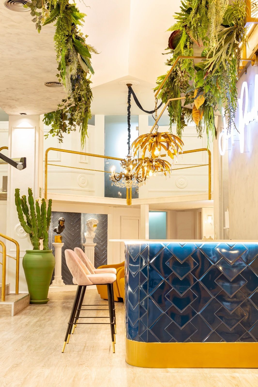 Barra con azulejos azules, detalles dorados y plantas colgantes; un taburete rosa.