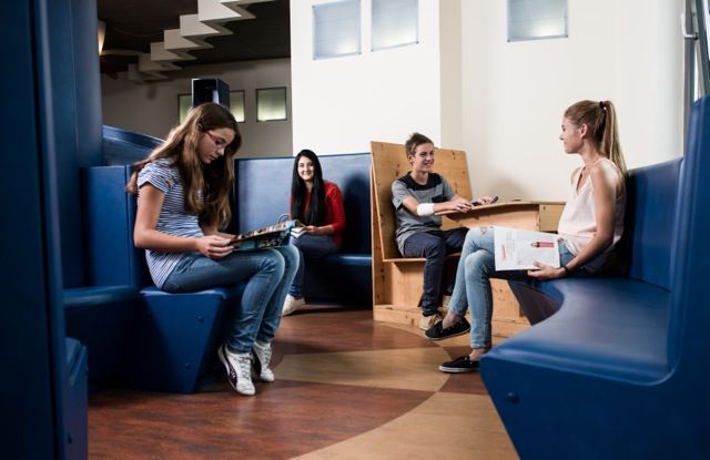 Studenten sitzen und unterhalten sich in einer modernen Lounge mit blauen Sitznischen und Holztischen.