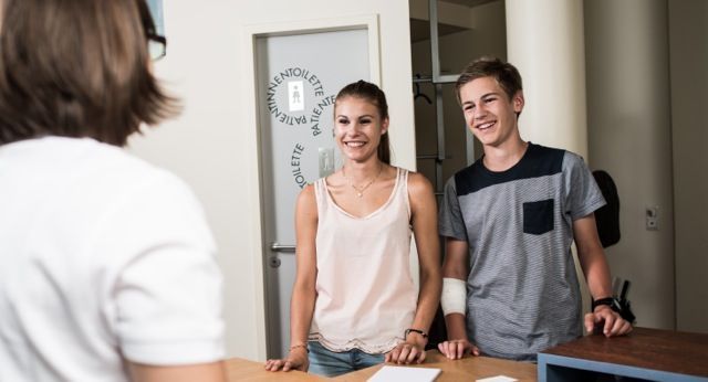 Zwei lächelnde Besucher stehen an einem Empfangstresen, einer von ihnen hat einen bandagierten Arm.