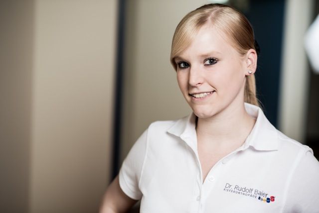 Eine lächelnde Person in einem weißen Poloshirt steht in einem Innenraum vor einem verschwommenen Hintergrund.