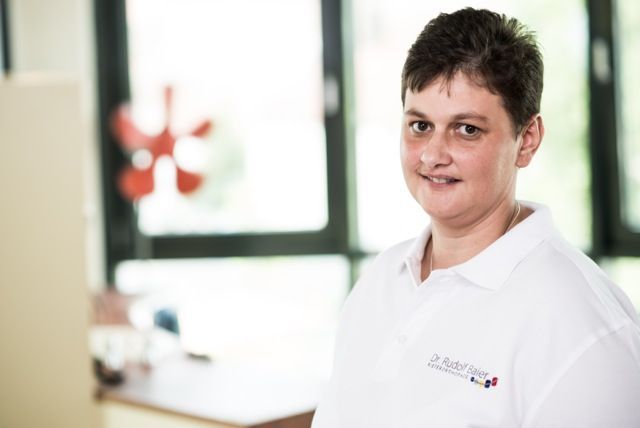Eine Person in einem weißen Poloshirt steht in einem Innenraum; im Hintergrund sind ein verschwommenes Fenster und eine rote Blumendekoration zu sehen.