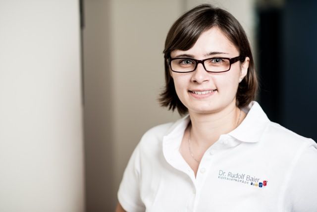 Eine lächelnde Person mit Brille und weißem Poloshirt steht drinnen vor einer schlichten Wand.