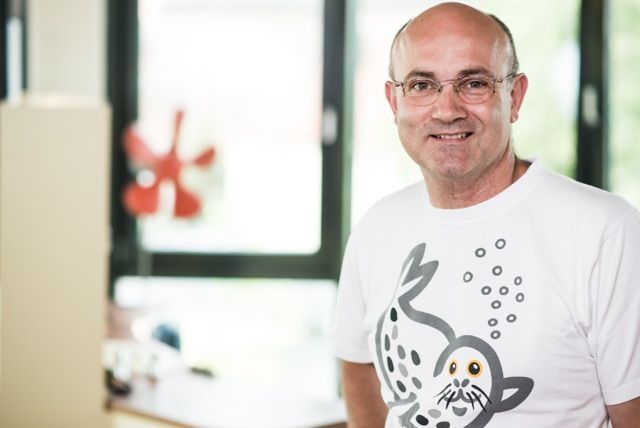 Eine lächelnde Person mit Brille und weißem Poloshirt steht drinnen vor einer schlichten Wand.