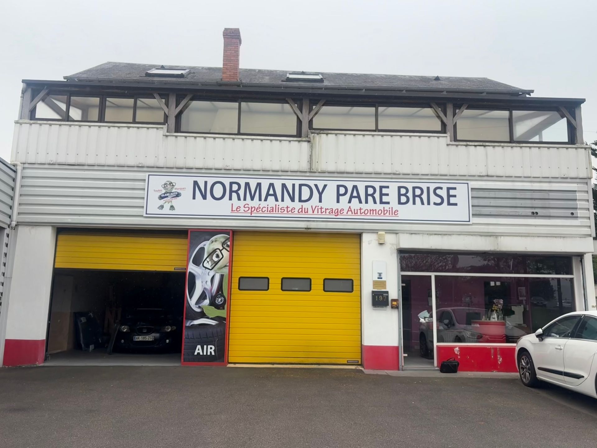Façade avec une enseigne NORMANDY PARE-BRISE, une porte et une voiture garée à l'extérieur.